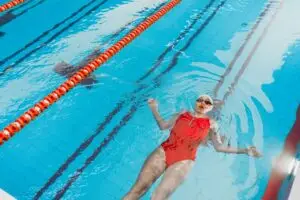 A woman in a red swimsuit floats on her back in an indoor swimming pool, enjoying a relaxing swim.